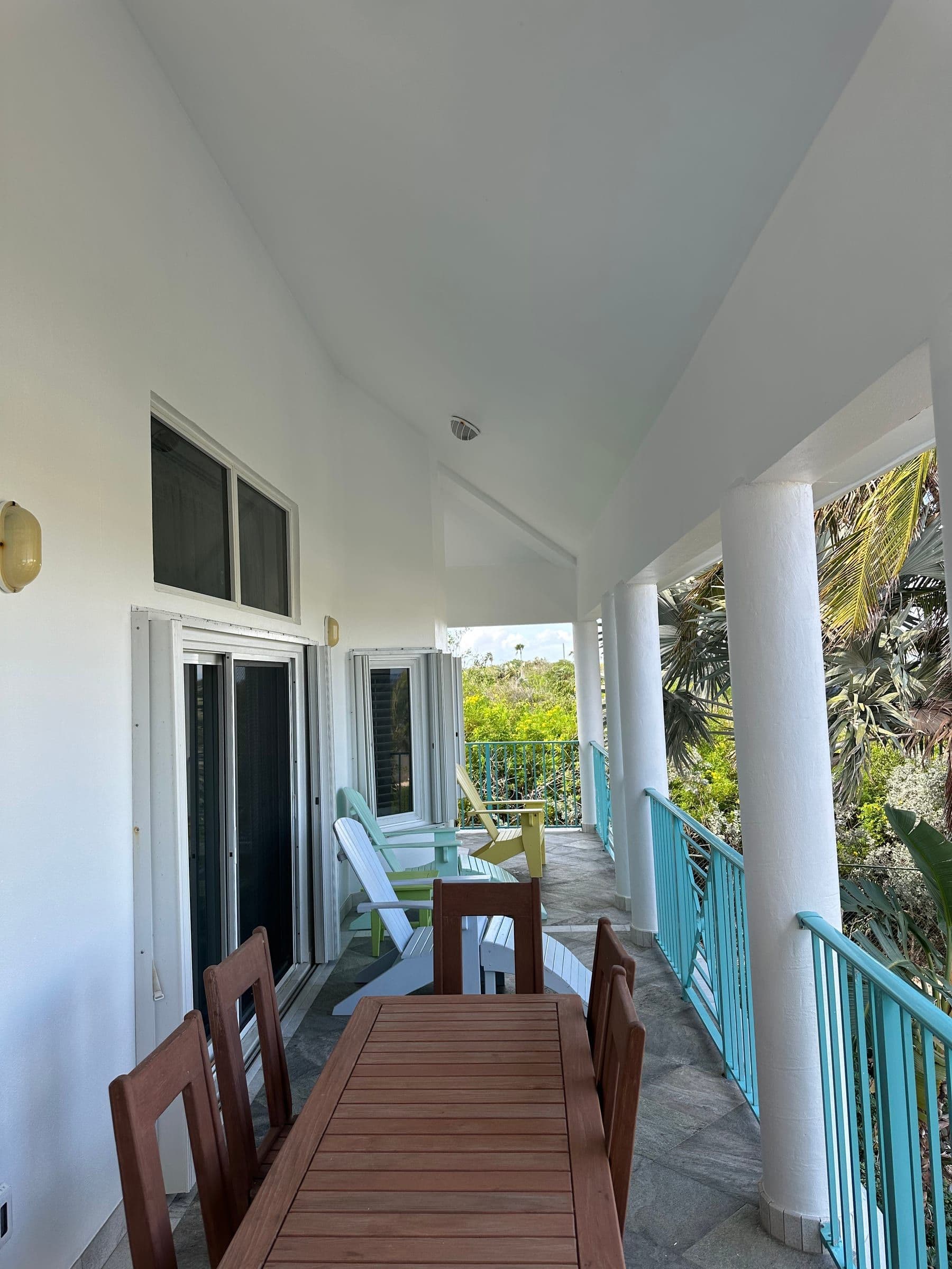The covered porch. Long wooden dining table, blue railings, palms beyond.